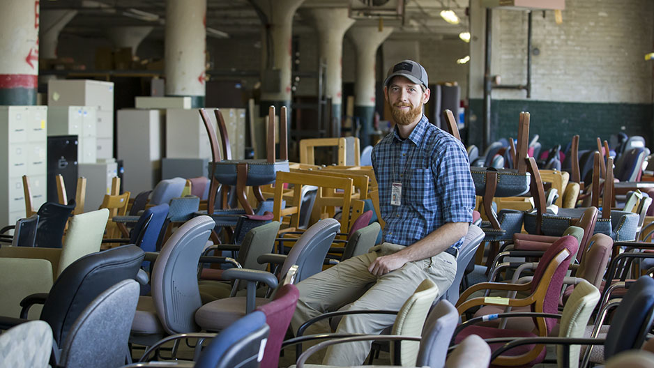 A man sitting amid cabinets, desks, chairs and other items in a warehouse.