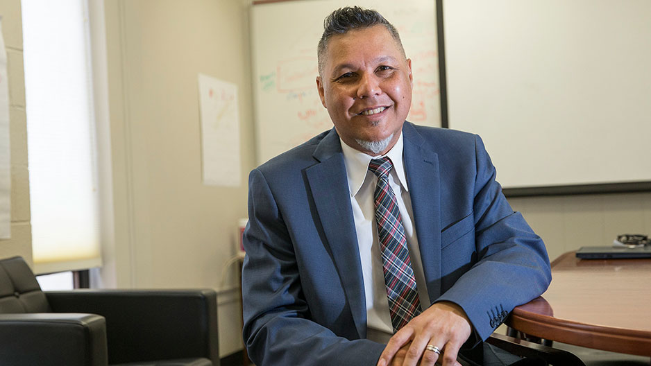 A man in a blue suit and plaid tie sits in a chair at a table.