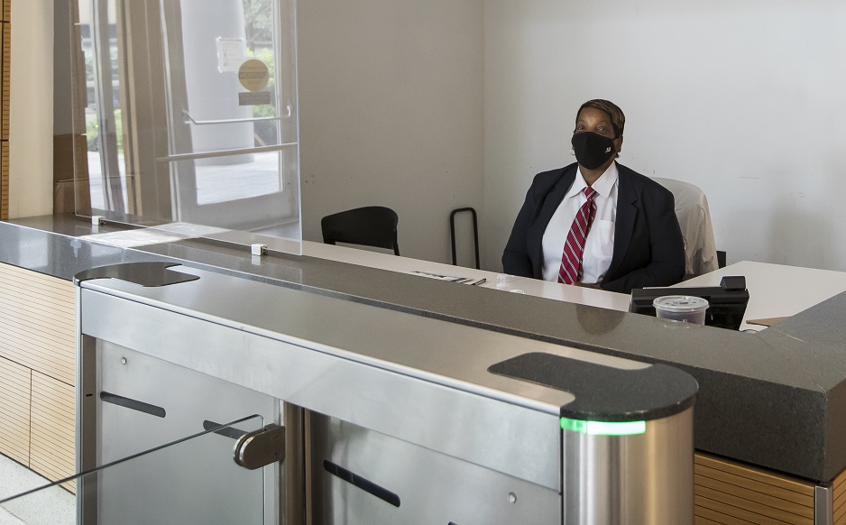 A security guard on campus wearing a mask and sitting behind a Plexiglas barrier at the entrance of a campus building.