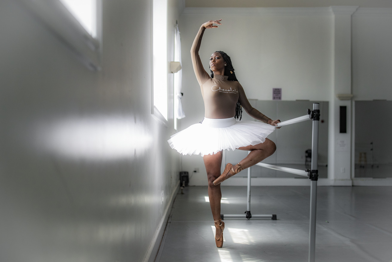 A Black woman in white tutu practicing ballet at a barre in a dance studio.