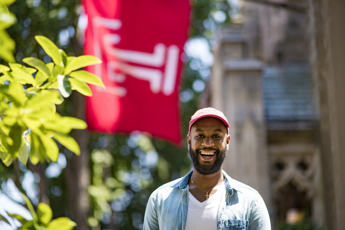 Brice Izyah smiling, wearing a red hat and standing under a Temple flag.