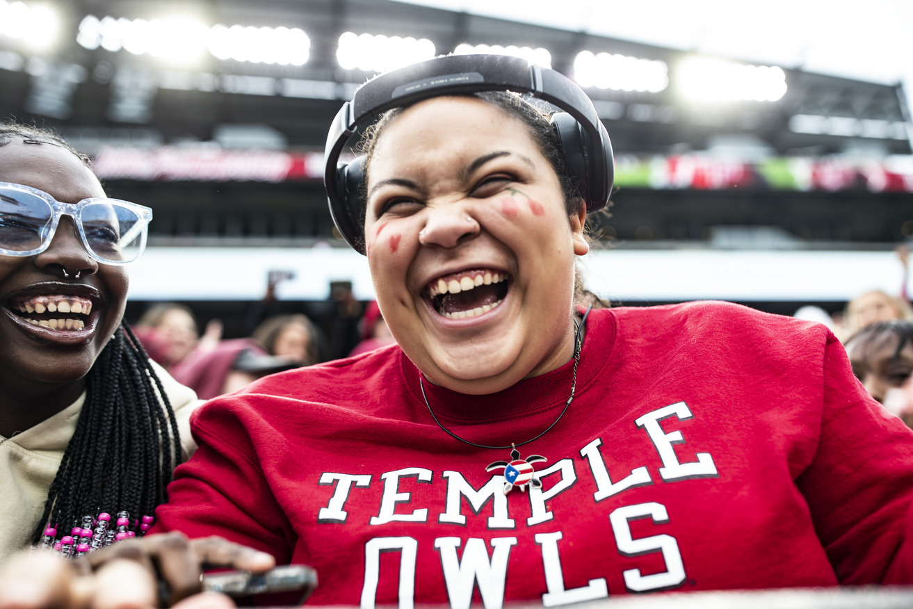 Image of a college student smiling, wearing a cherry and white shirt, seated at a Temple University homecoming event.