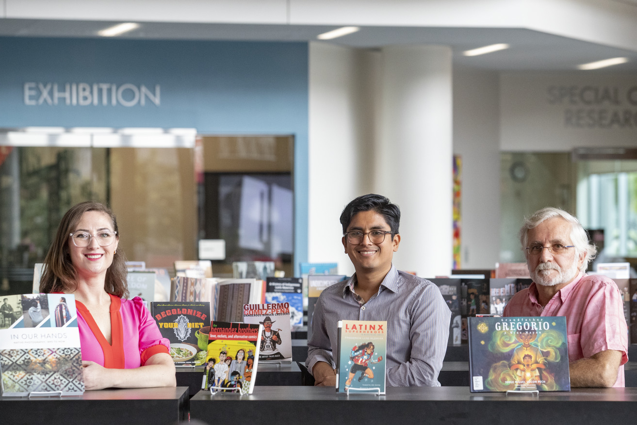 Professors Christina Baker, C sar Cabezas and Augusto Lorenzino standing in front of books on display in Charles Library for Hispanic Heritage Month