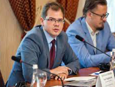 image of Ivan wearing glasses, in a suit sitting at a table speaking into a microphone
