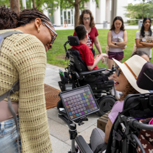 Students using AAC devices and wheelchairs mingle and chat outdoors on Temple's campus with others
