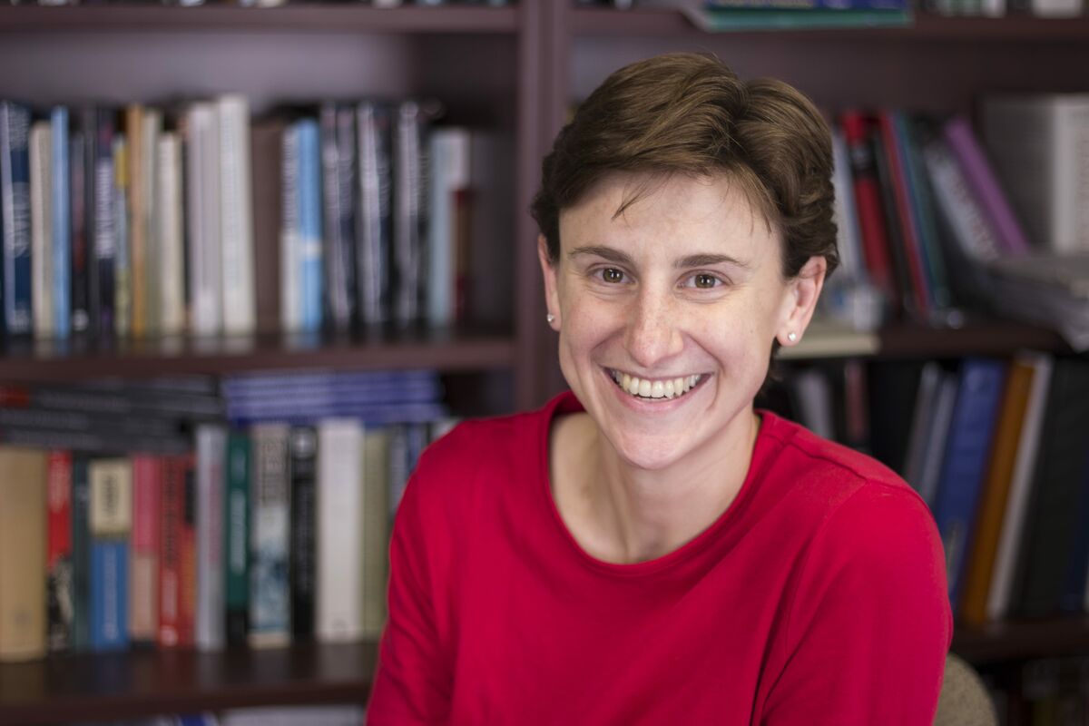 This photo is a headshot of Dr. Kate Epstein. She is smiling and wearing a red shirt in front of a bookcase.