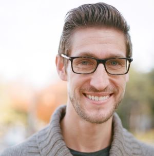 image of Jacob standing outside wearing glasses and a tan sweater smiling at the camera