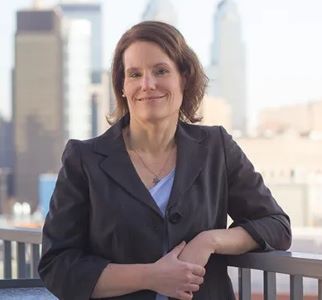 image of Teresa standing with the city in the background wearing a grey suit and blue top with her arms crossed