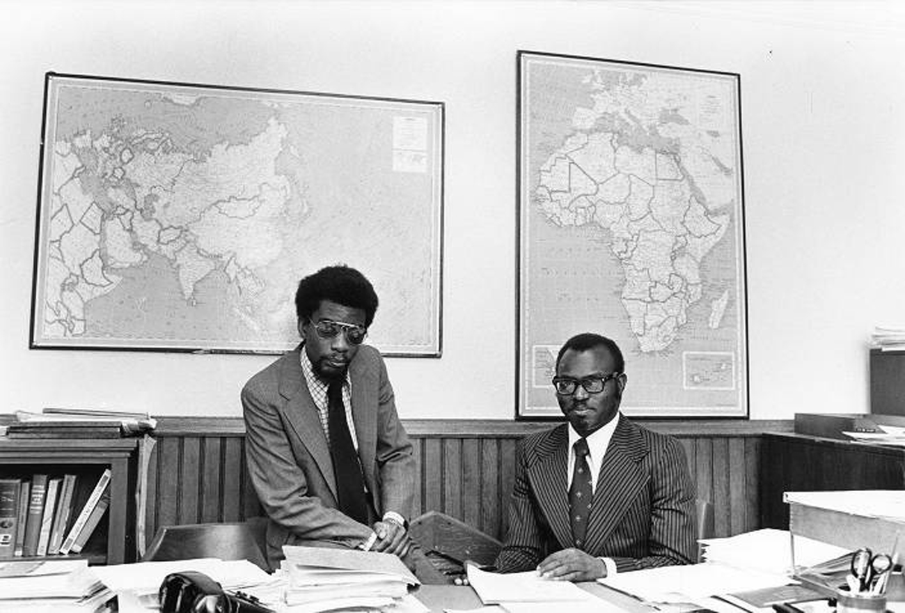 two men wearing suits sitting at a desk.