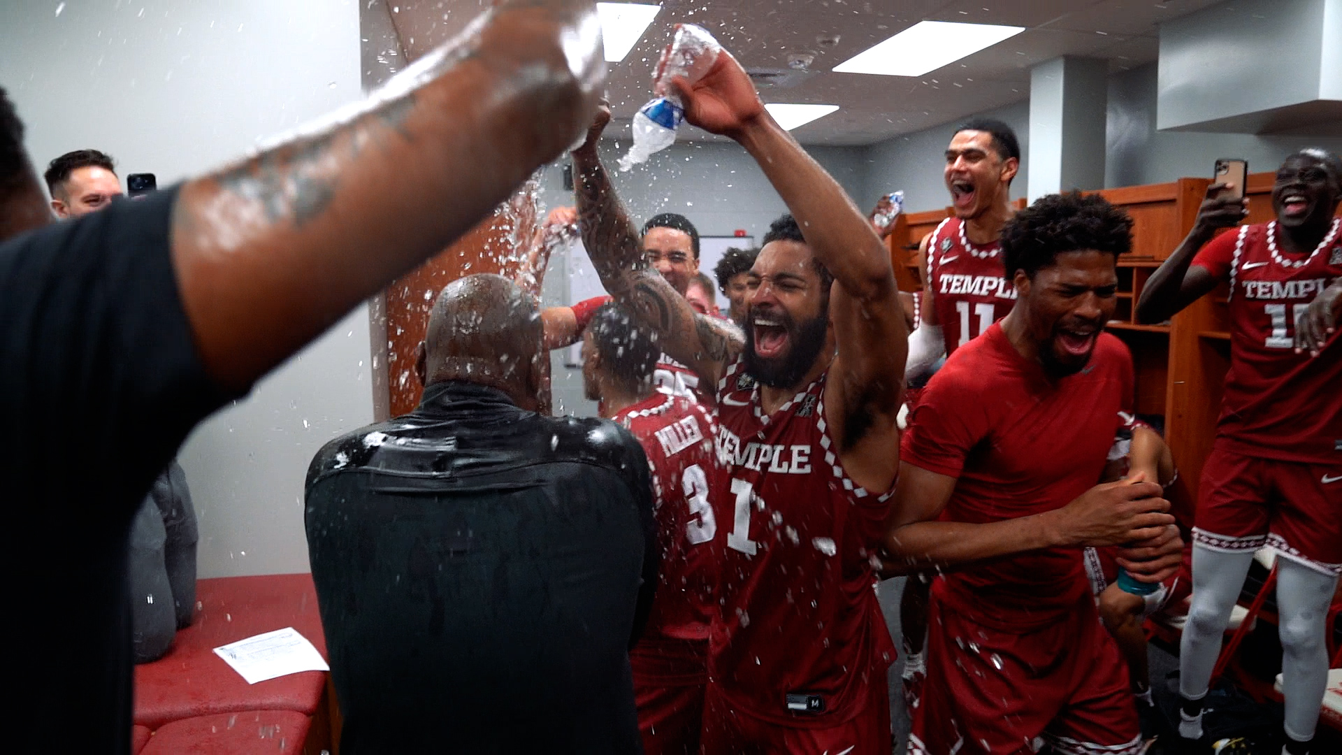 Temple basketball players wearing cherry and white jerseys, smiling brightly douse water on a coach inside the team locker room.