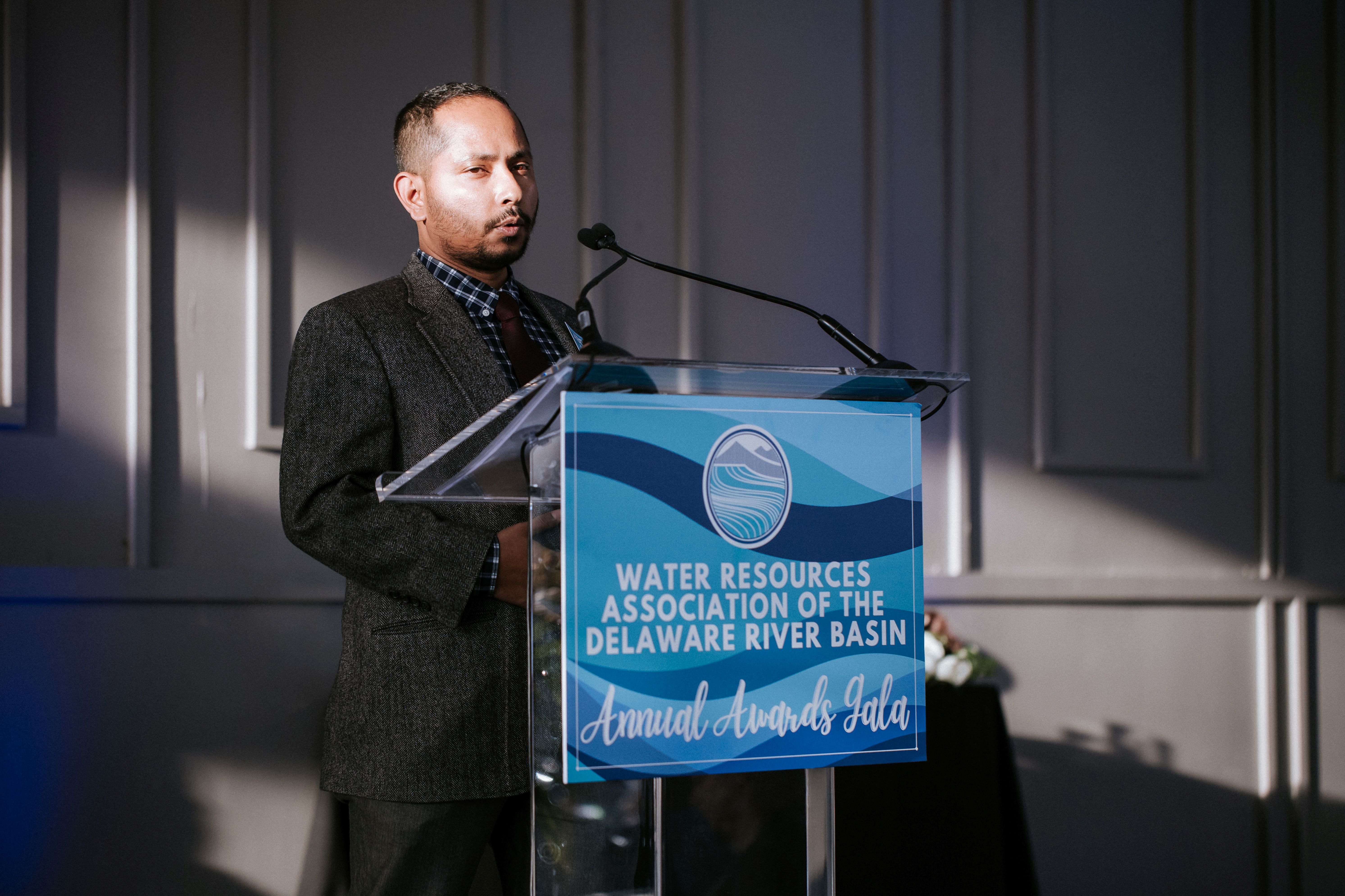 A man in a suit and tie stands speaking at a podium during an awards gala.
