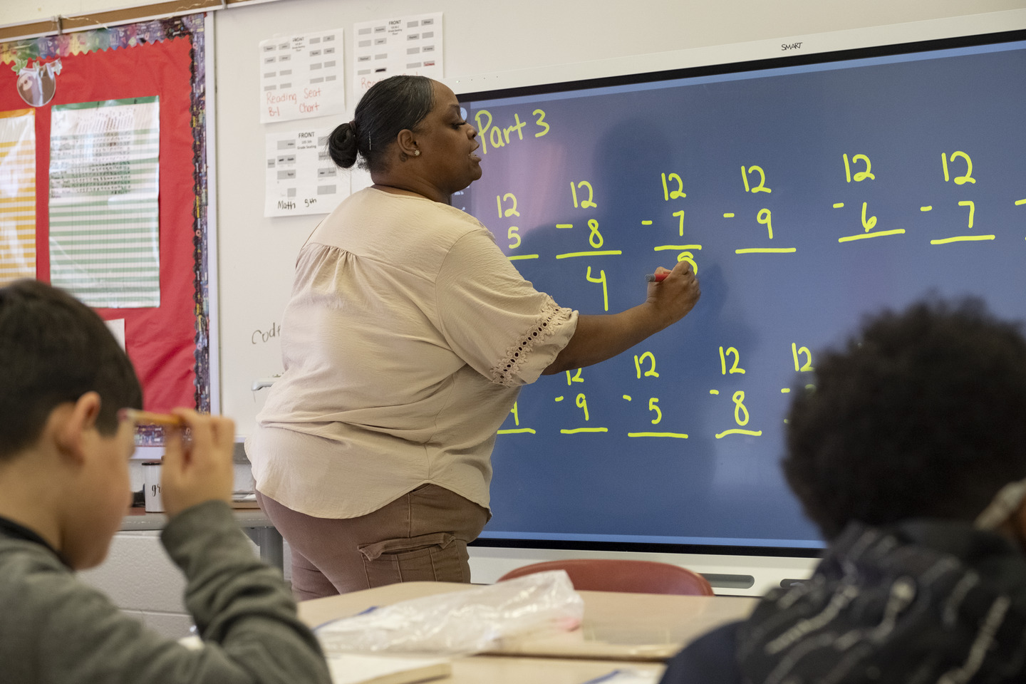 A Black female teacher demonstrates subtraction on a screen in an elementary school classroom with students looking on