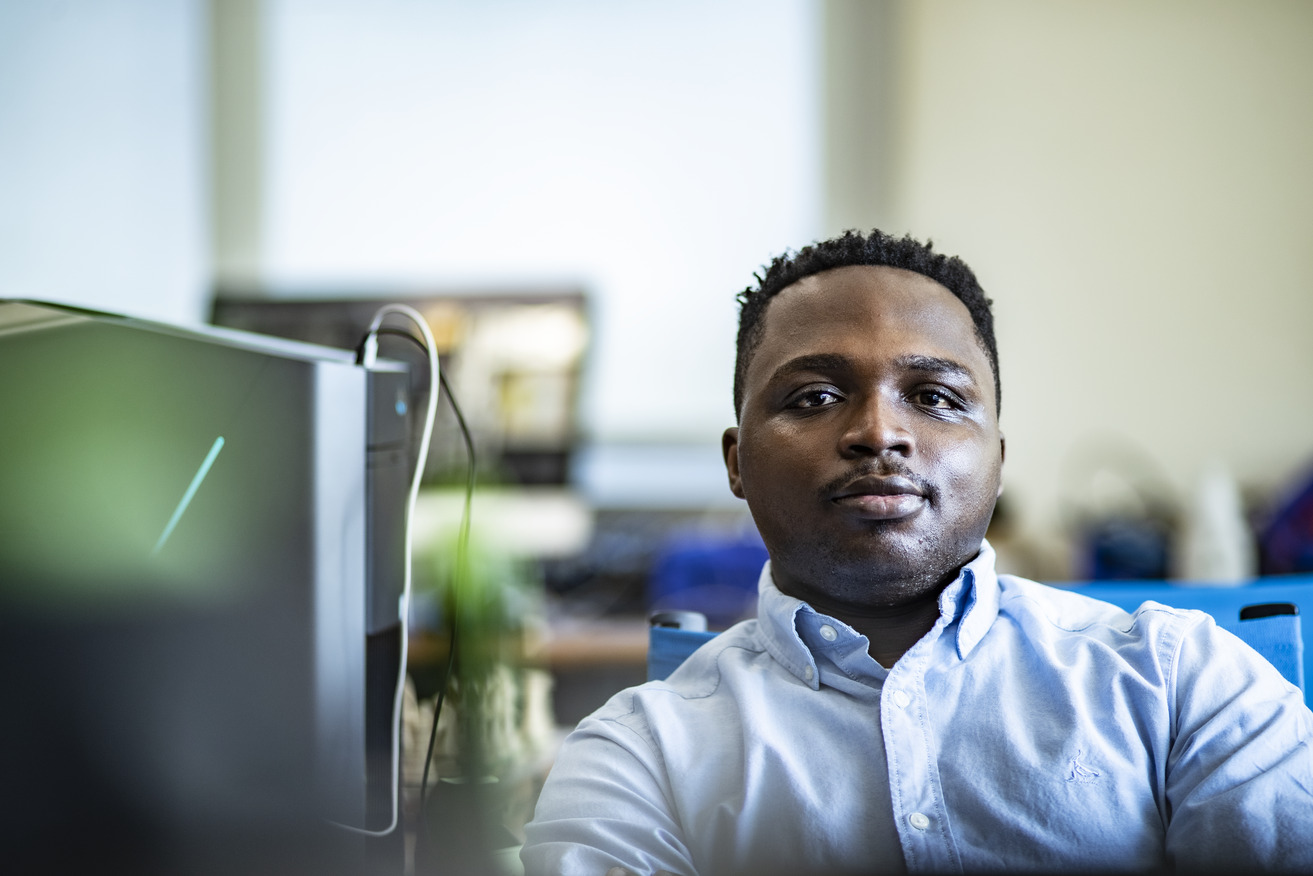 A student sits in a computer lab. He is wearing a button down shirt and looking forward.