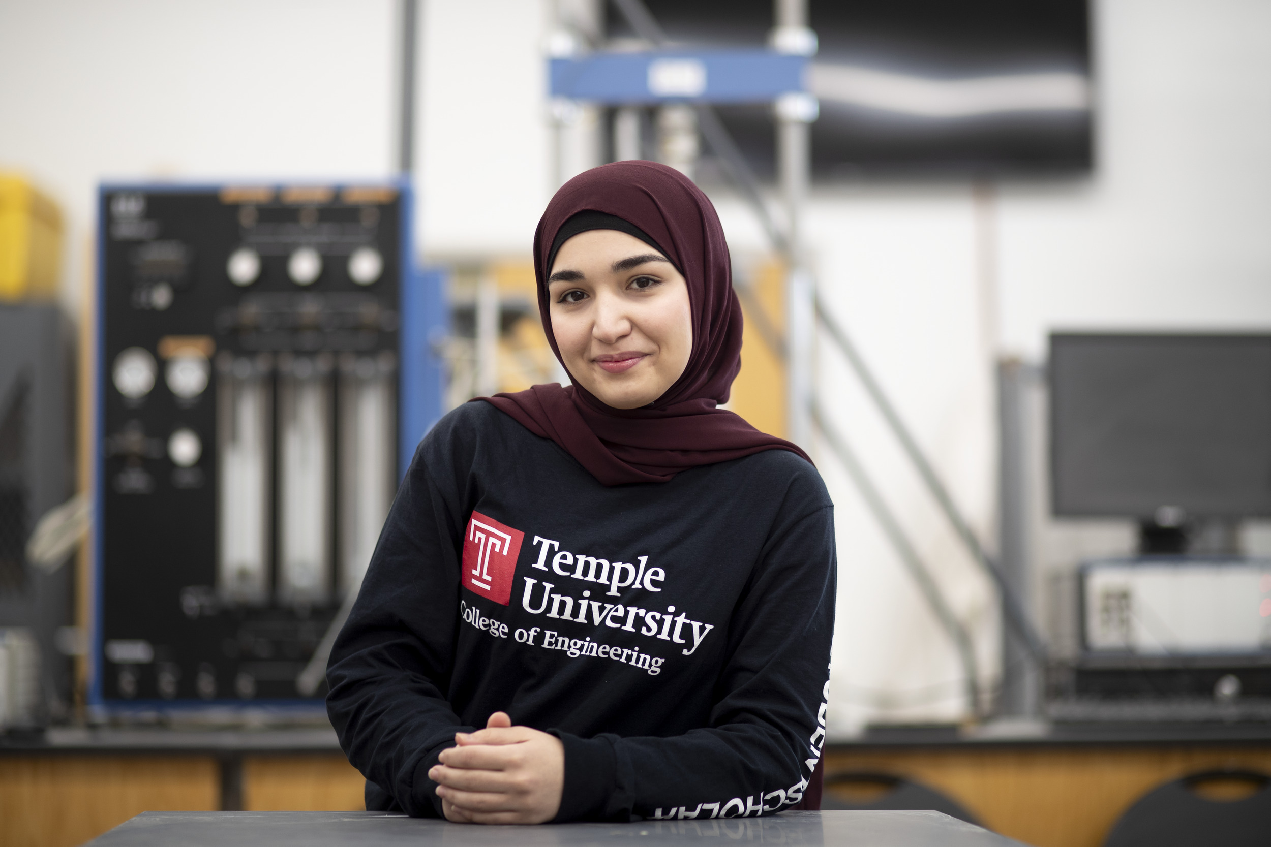 A student stands at a lab table, smiling with her hands folded. There is various lab equipment in the background.