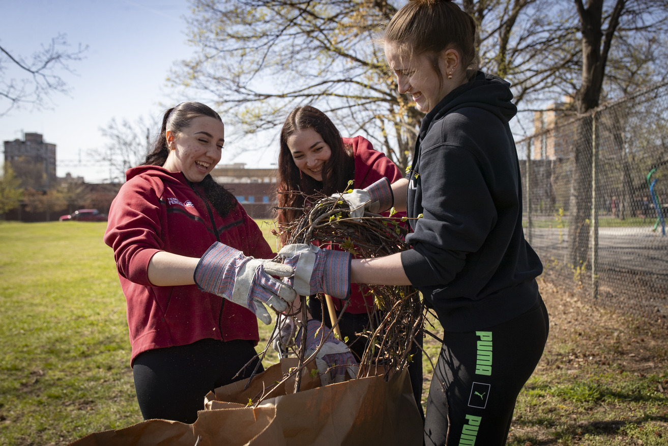 Three young women wearing gloves smile and work together outdoors, gathering sticks and debris into a bag in a park on a sunny day.