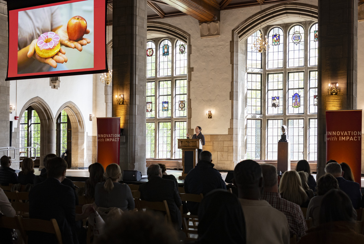 Image of a speaker standing at a podium in a cathedral-like hall, addressing an audience.. A large screen to the left shows a close-up of hands holding an apple and a decorated donu