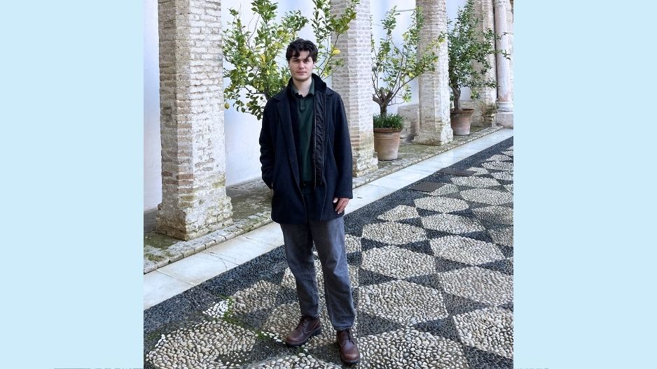 Aidan stands on a stone walkway with arches and greenery behind him
