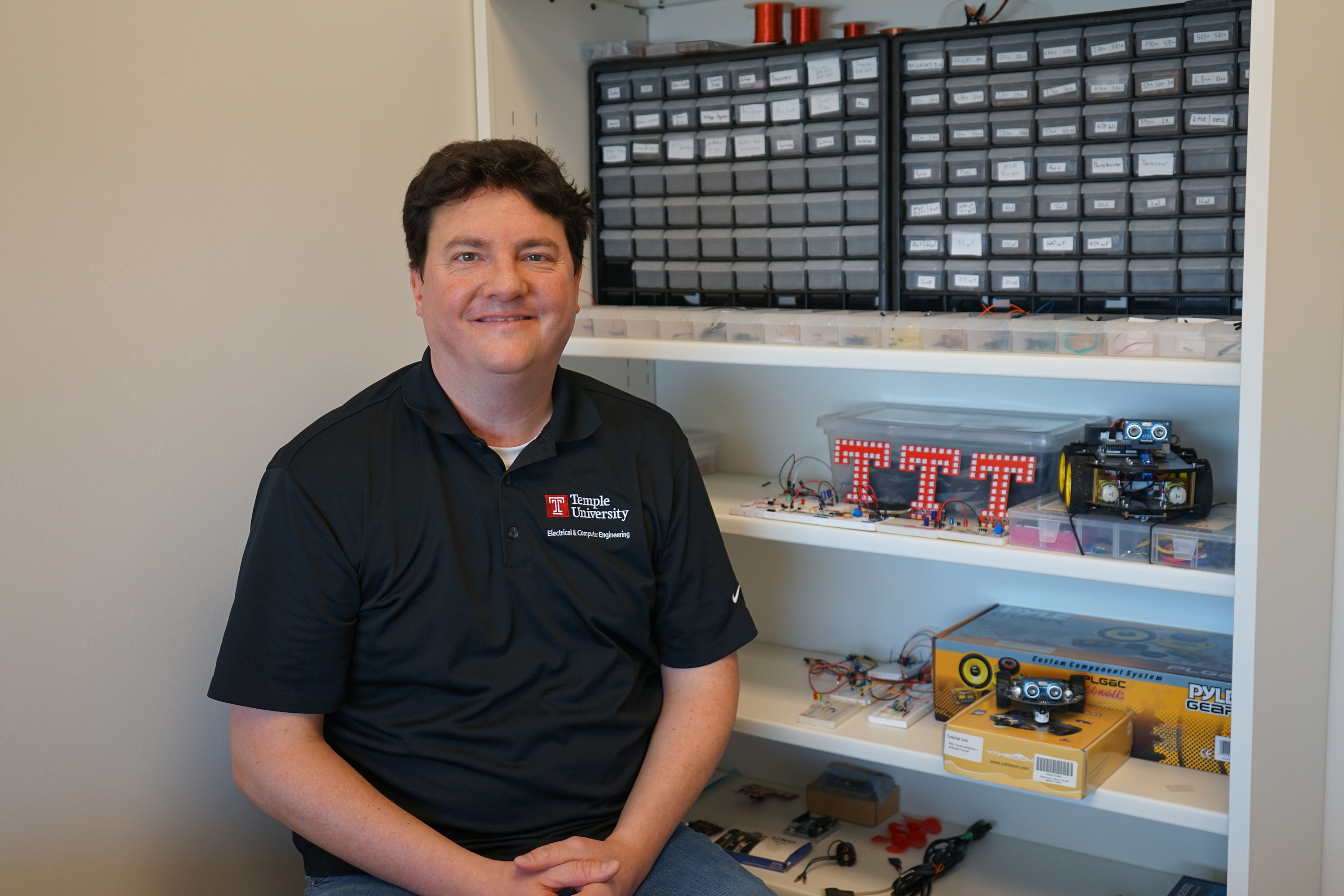 A faculty member seated to the side of a bookshelf, smiling. The bookshelf has various electrical engineering hardware.