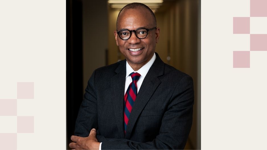 James Davis poses for a headshot in a dimly lit hallway. He is a Black man wearing eyeglasses, a dark suit jacket, white shirt, and red and blue tie.