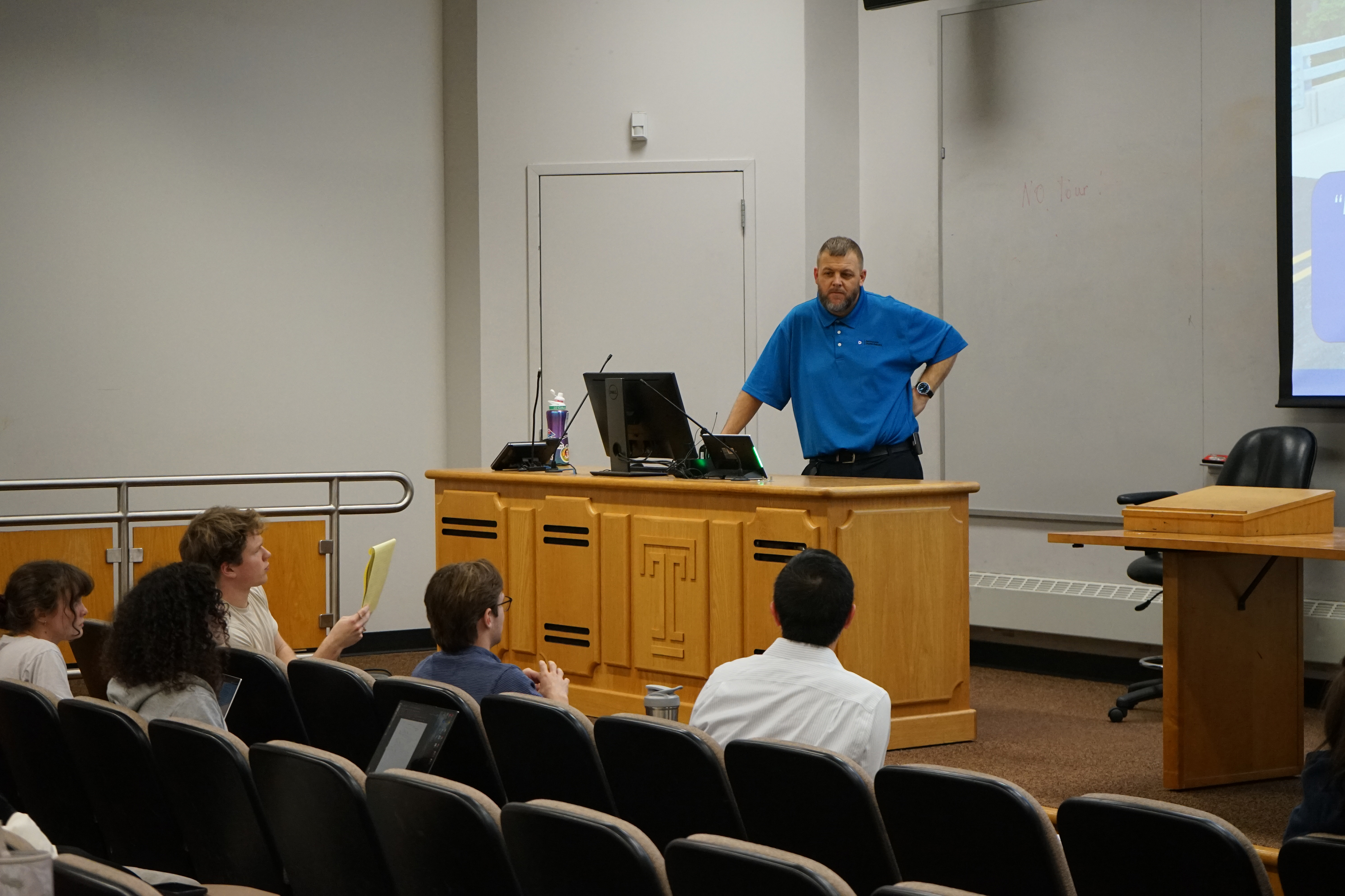 A lecturer stands at the front of a classroom. He is leaning on the desk and listening to a student ask a question.