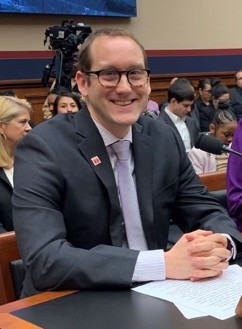 Man with glasses wearing grey suit, white shirt sitting at a table at a lecture