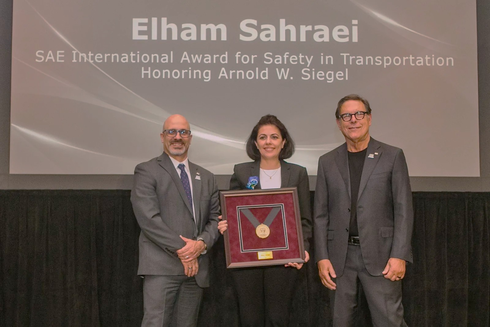 Elham Sahraei stands on stage holding an award. Jay Solomond and Robert Ireland stand on either side of her.