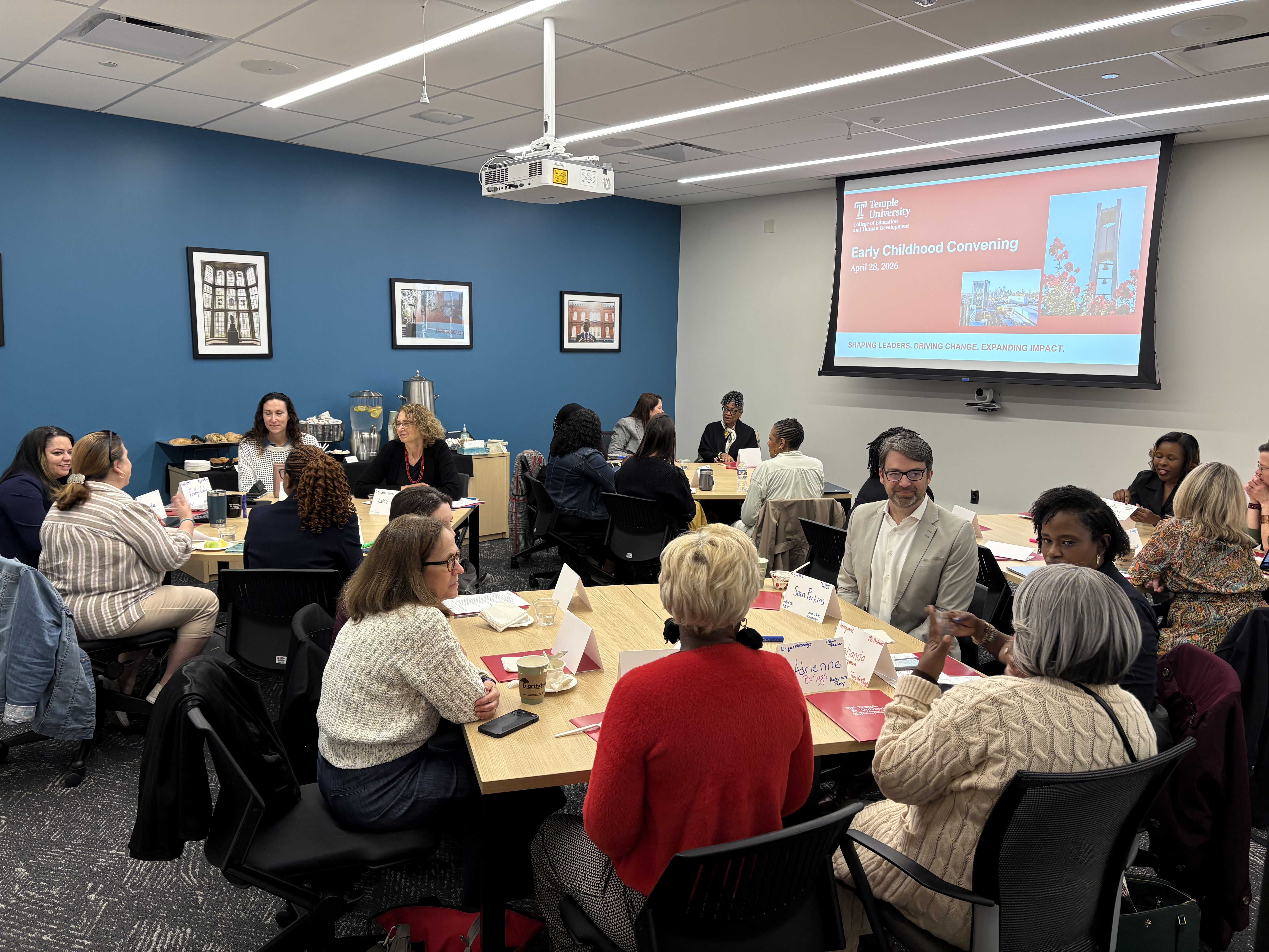 A room full of people engaged in workgroups at table pods with a presentation on display in the background