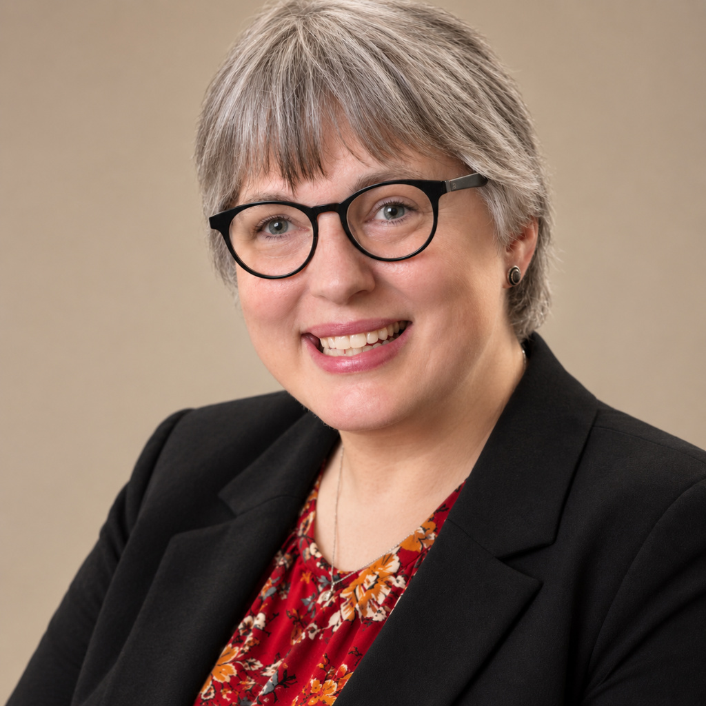 Jamie Ray-Leonetti, a smiling white woman with short salt and pepper hair, wears eyeglasses and business attire