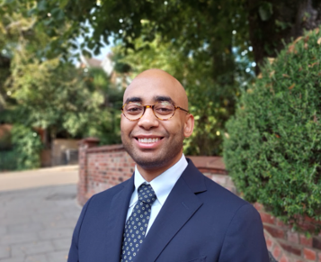 Man with wire glasses wearing blue suit, light blue shirt and blue tie in a park 