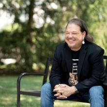 image of Jeremy Johnson sitting on a bench wearing a black blazer, jeans and is smiling looking into the distance