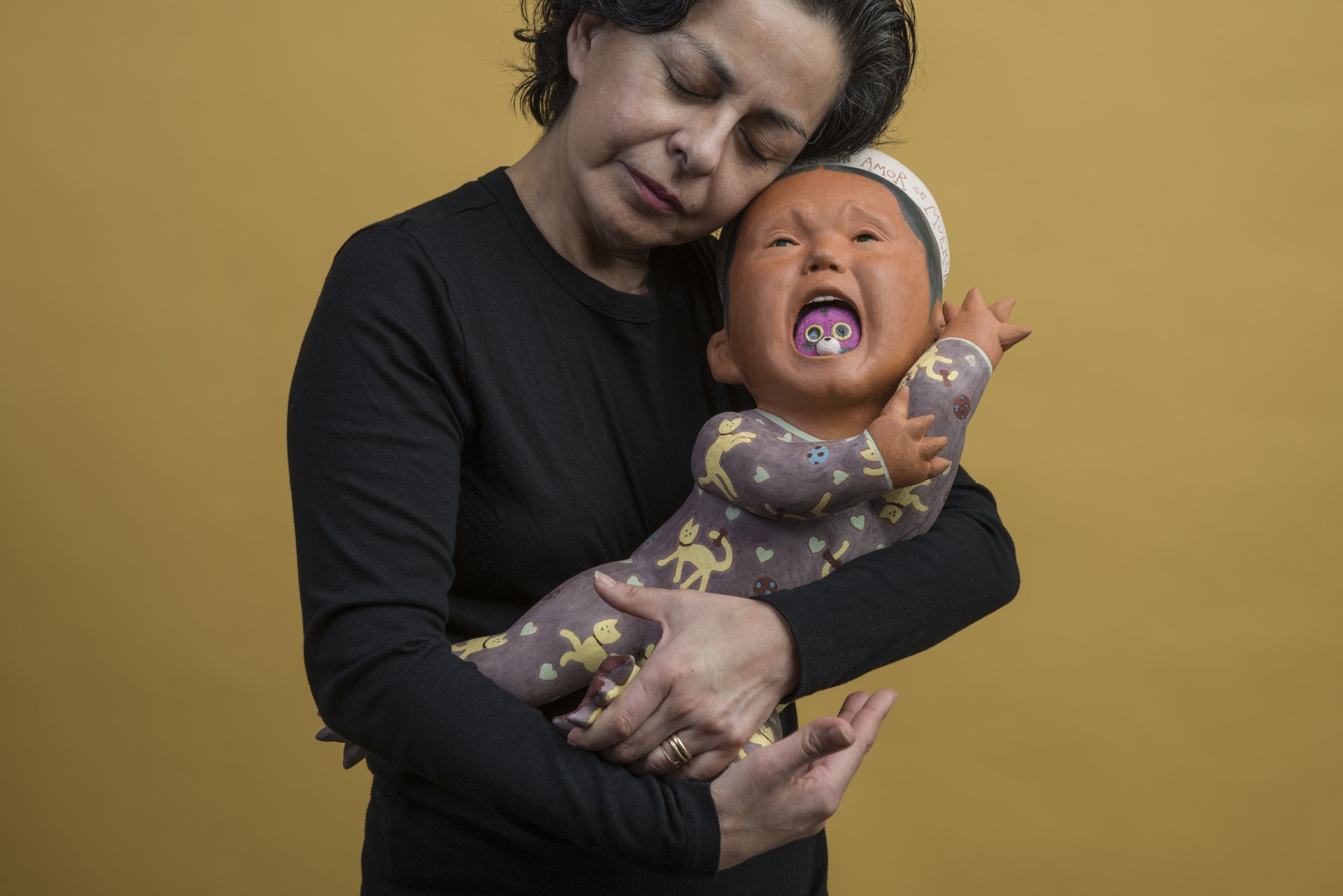 Woman holding Baby clay sculpture that appears to be crying has purple round object in mouth