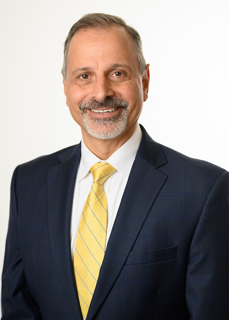 A man in a suit and yellow tie is looking forward and smiling in front of a plain white background.