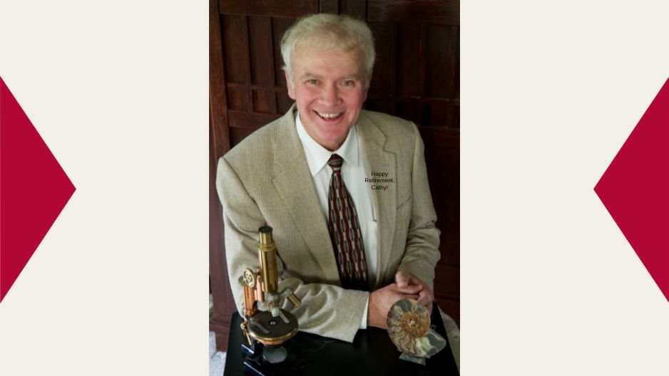 Dr. George Mehler sits in a tan suit with his arms folded leaning slightly on a table that has a microscope on it
