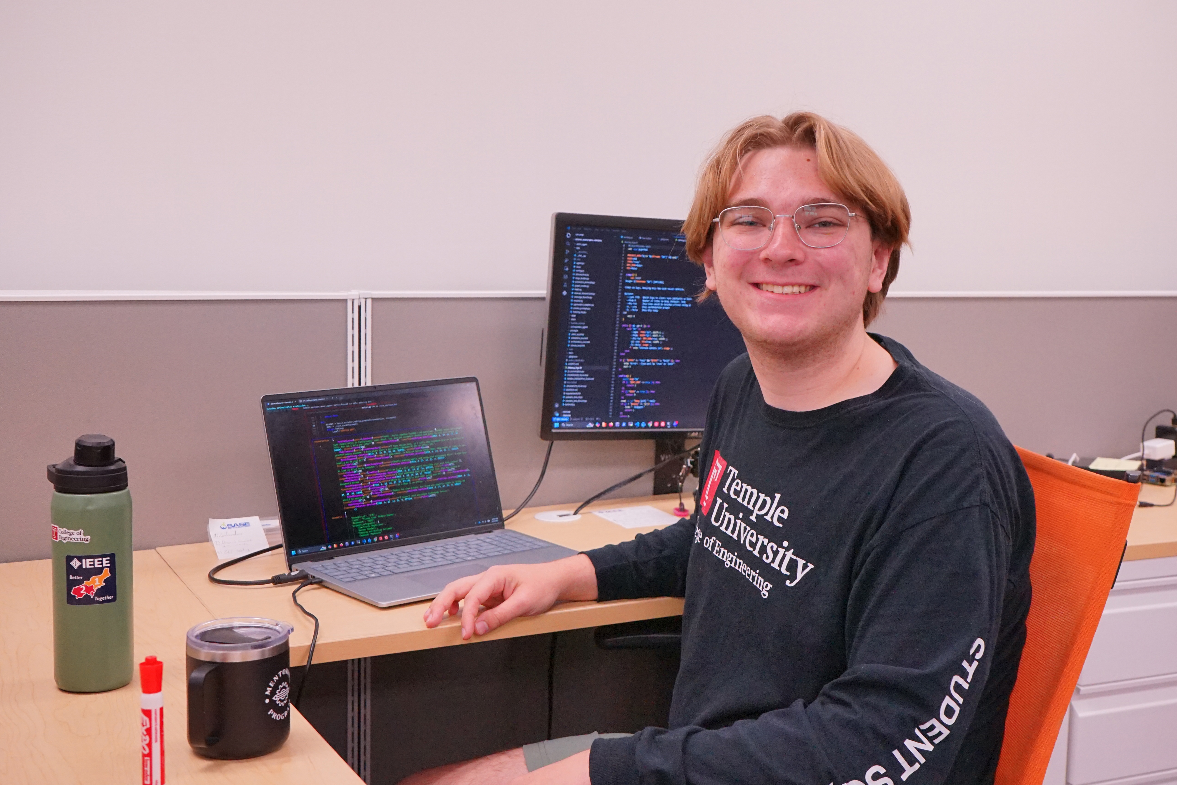 A student sits at a desk with a laptop and monitor displaying code. He is looking forward and smiling.