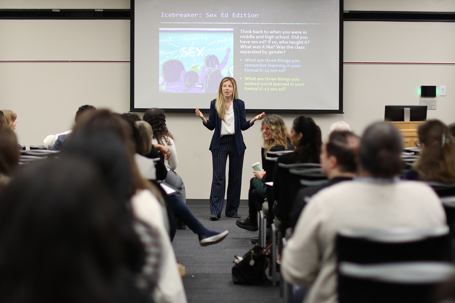 A woman stands at the front of a full classroom sharing a presentation
