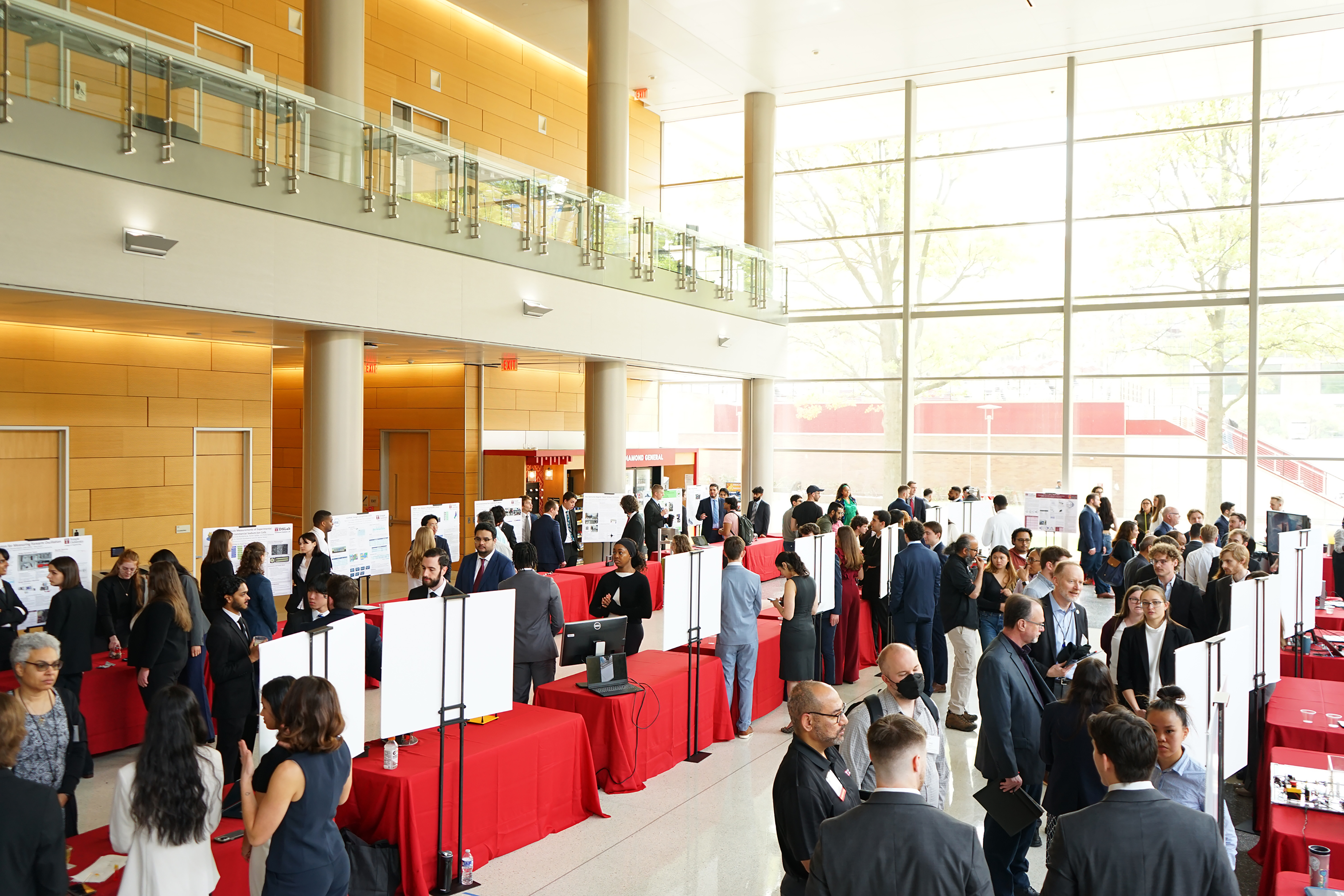Several tables and poster stands are set up in a crowded lobby. People are viewing each poster and speaking with one another.