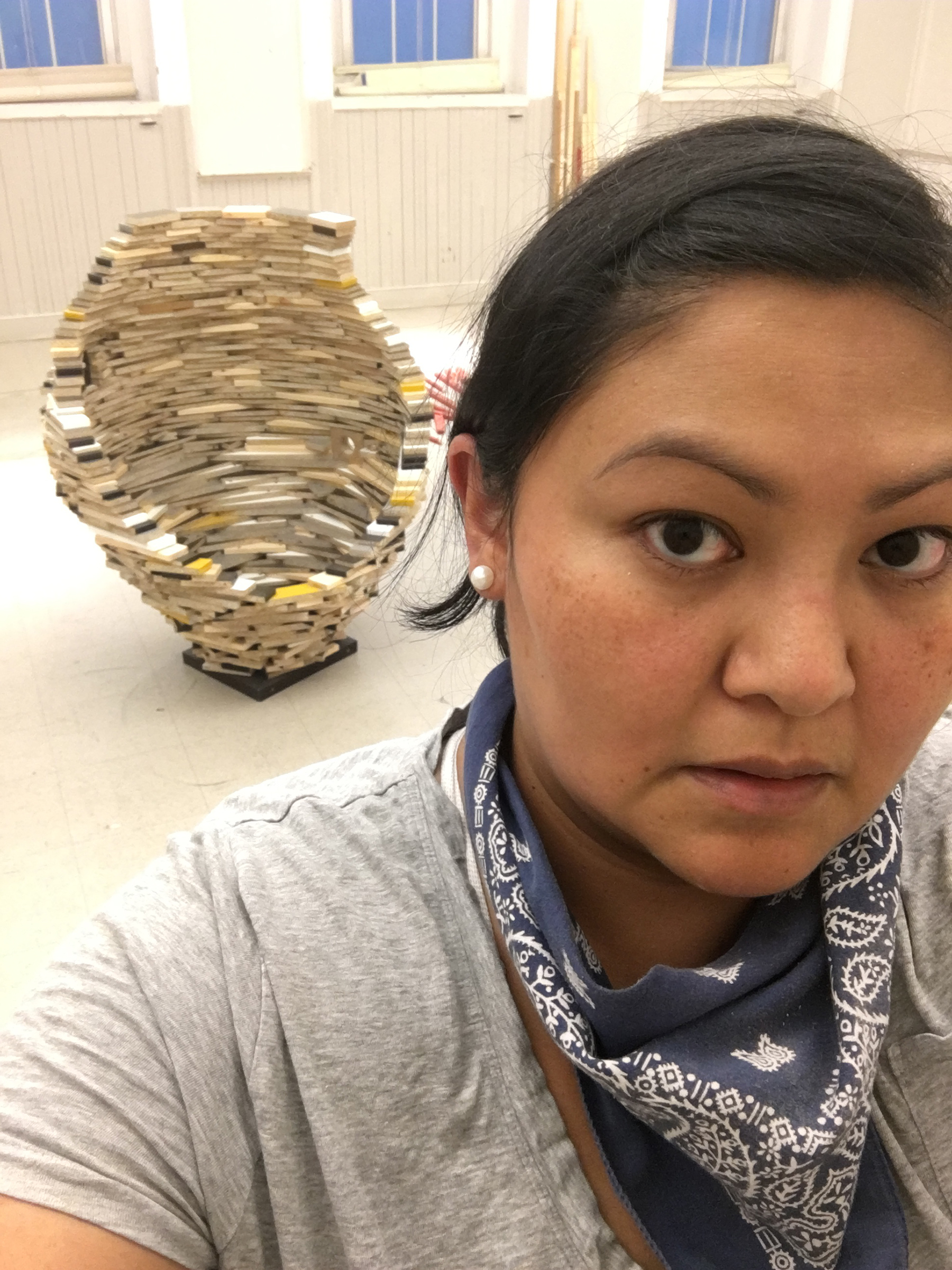 Anna Tsouhlarakis, a person with dark hair and eyes, looks at the camera. Behind her is a large sculpture in her studio. She wears a gray shirt and a blue bandana around her neck.