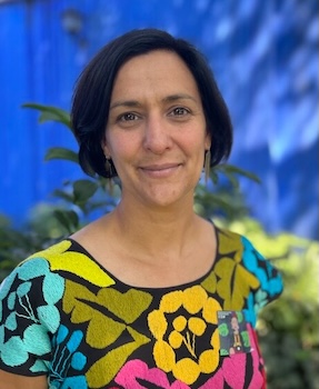 headshot of Caroline Faria in a floral blouse standing in front of a garden and blue wall