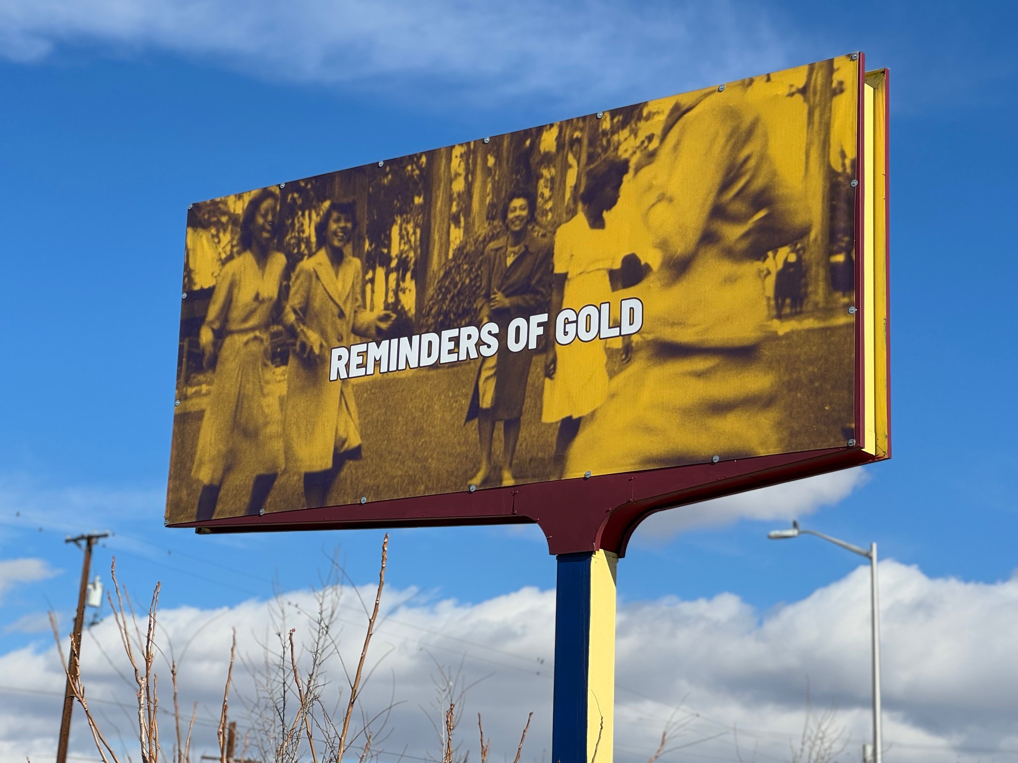 A billboard with a gold-tinted photograph of people is overlaid with text reading "Reminders of Gold" in front of a blue sky.