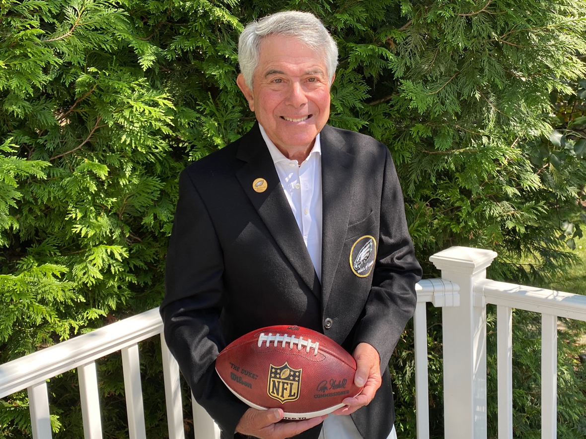 Merrill Reese holding a football and wearing his Eagles Hall of Fame jacket outside.