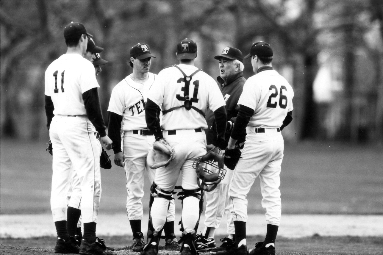 An older man with a group of student-athletes wearing caps, in black and white uniforms, standing on a raised dirt area at a Temple baseball game.
