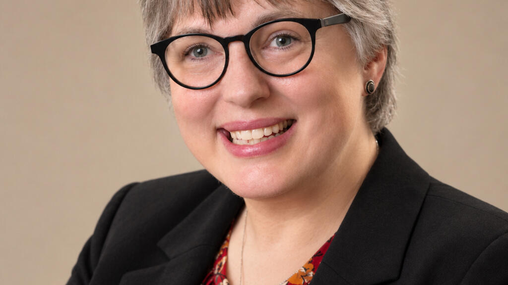 Jamie Ray-Leonetti, a smiling white woman with short salt and pepper hair, wears eyeglasses and business attire