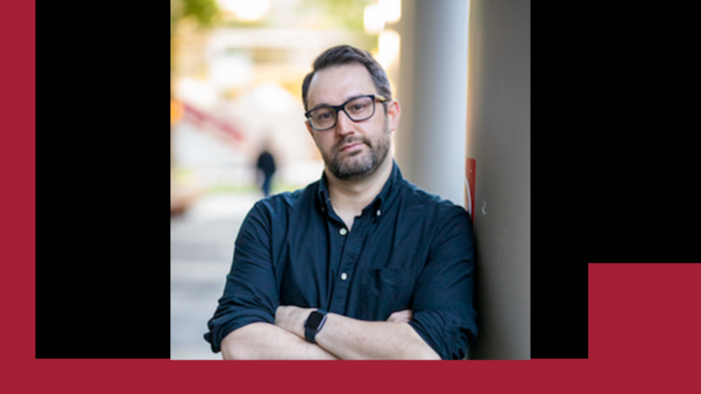 image of Jason standing with his arms crossed leaning against a wall wearing a blue shirt and glasses