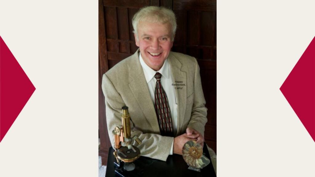Dr. George Mehler sits in a tan suit with his arms folded leaning slightly on a table that has a microscope on it