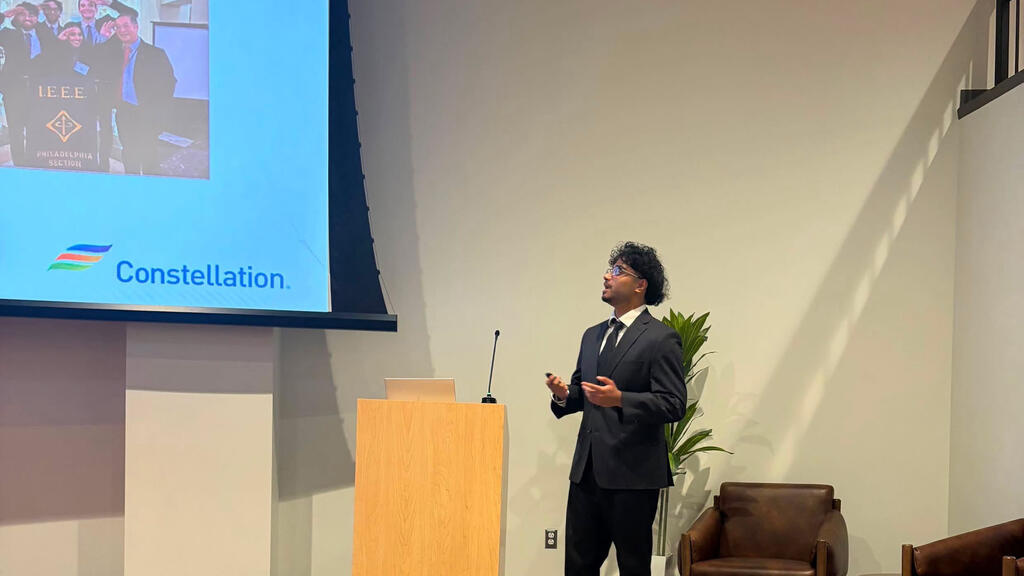 A student stands in front of a podium giving a presentation. He is wearing a suit and tie.