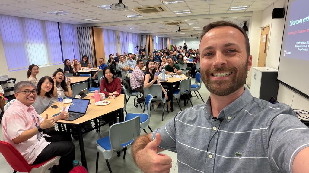 A man wearing a buttoned shirt takes a selfie with a clasroom full of educators sitting at tables