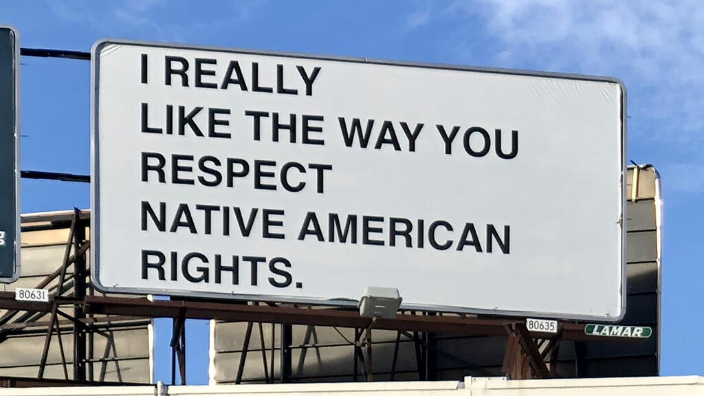 A billboard sits atop a building in a blue sky. In black letters on white background, the billboard reads "I REALLY LIKE THE WAY YOU RESPECT NATIVE AMERICAN RIGHTS."