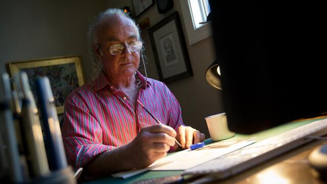 A man in a striped shirt drawing on paper at a desk.