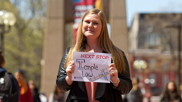 A woman in a black jacket holding a sign in front of Temple s bell tower.