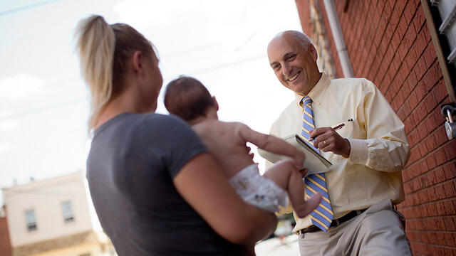 A man holding a notebook interviewing a woman holding a baby.